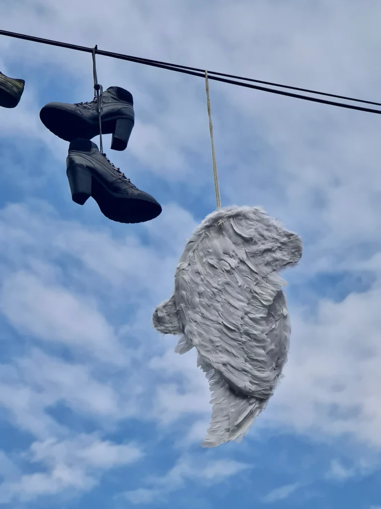 Zoomed-in image taken from below, showing detailed wings hanging from electrical cables, along with several pairs of shoes also suspended. A blue sky with clouds is visible in the background.