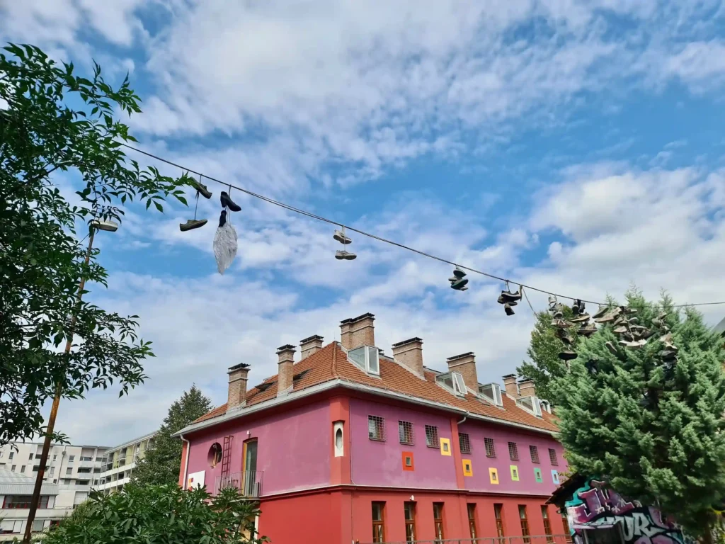 Wide image of buildings surrounding an art installation, with a two-story building in the background, trees framing the scene, and a cable with shoes and wings in the center, set against a blue sky with clouds