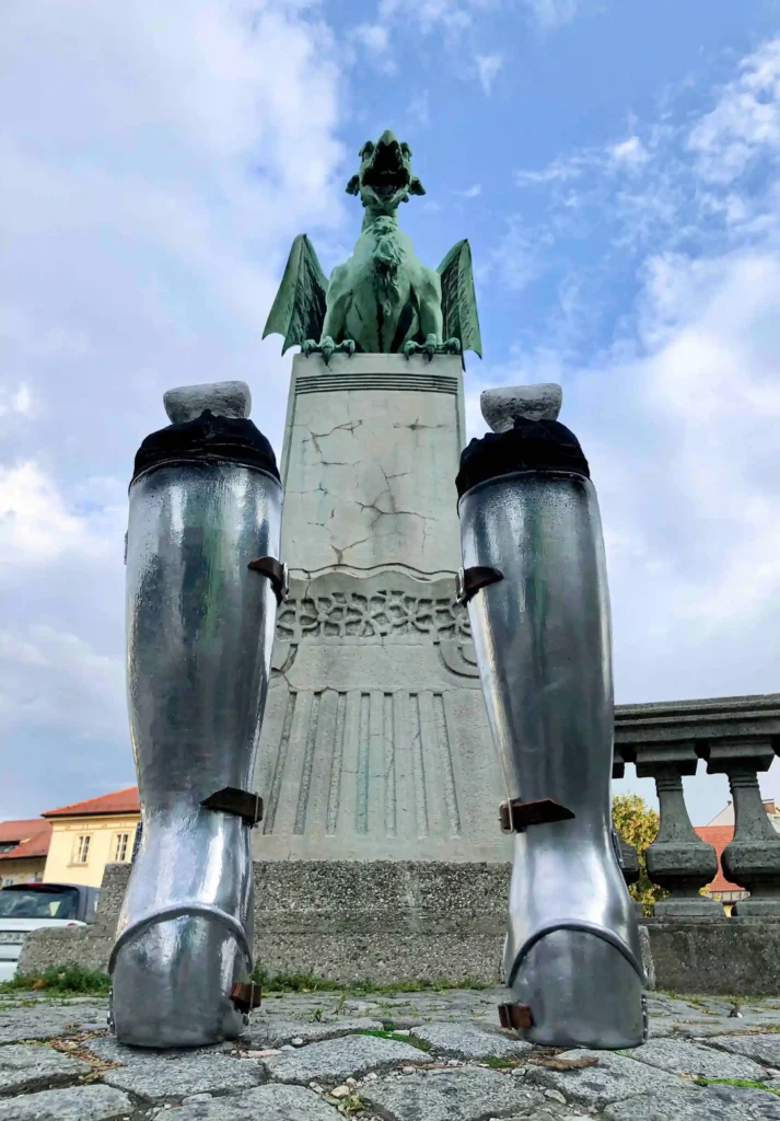 Half-burned medieval boots in front of a dragon statue on Dragon Bridge in Ljubljana, with a burned knight’s silhouette on the ground, viewed from a low angle, making it appear as if the dragon's fire burned the knight.