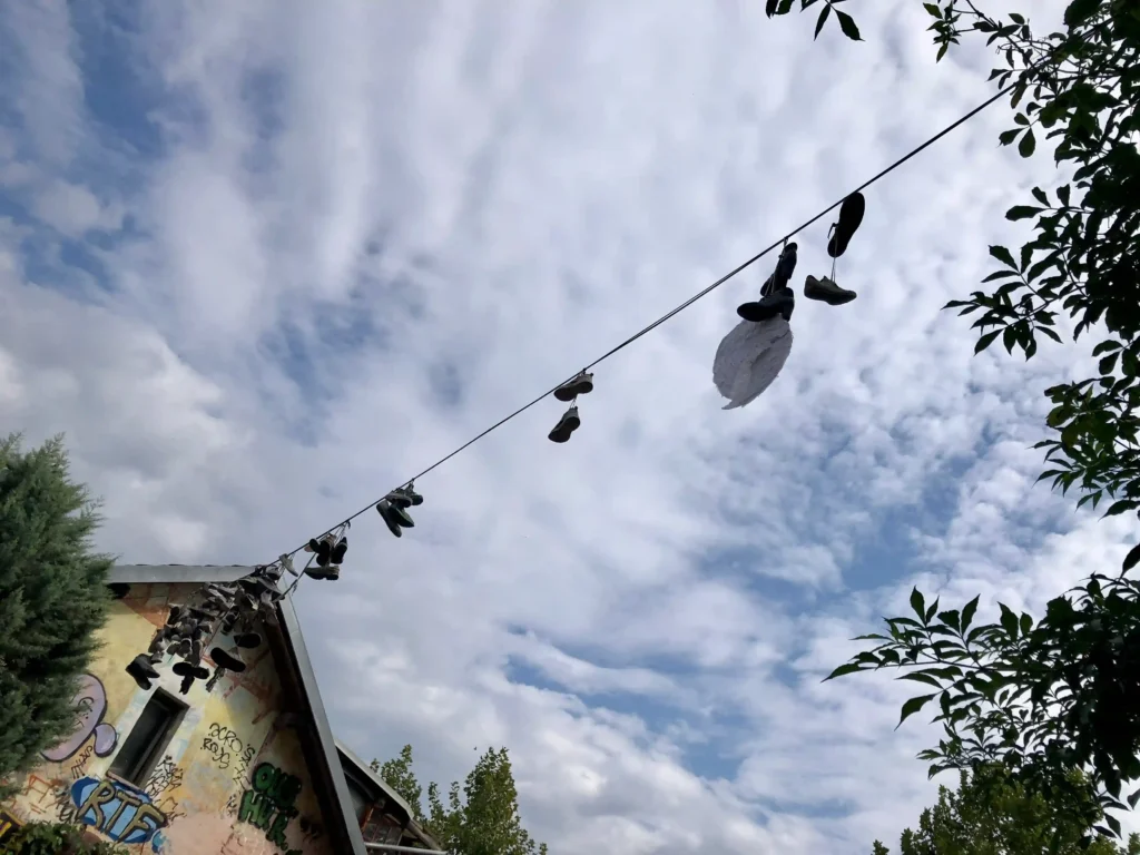 Wide image from the opposite side of the cable, displaying the building anchored with the cable that holds shoes and wings. The building is covered in graffiti, contrasted by a blue sky with clouds.