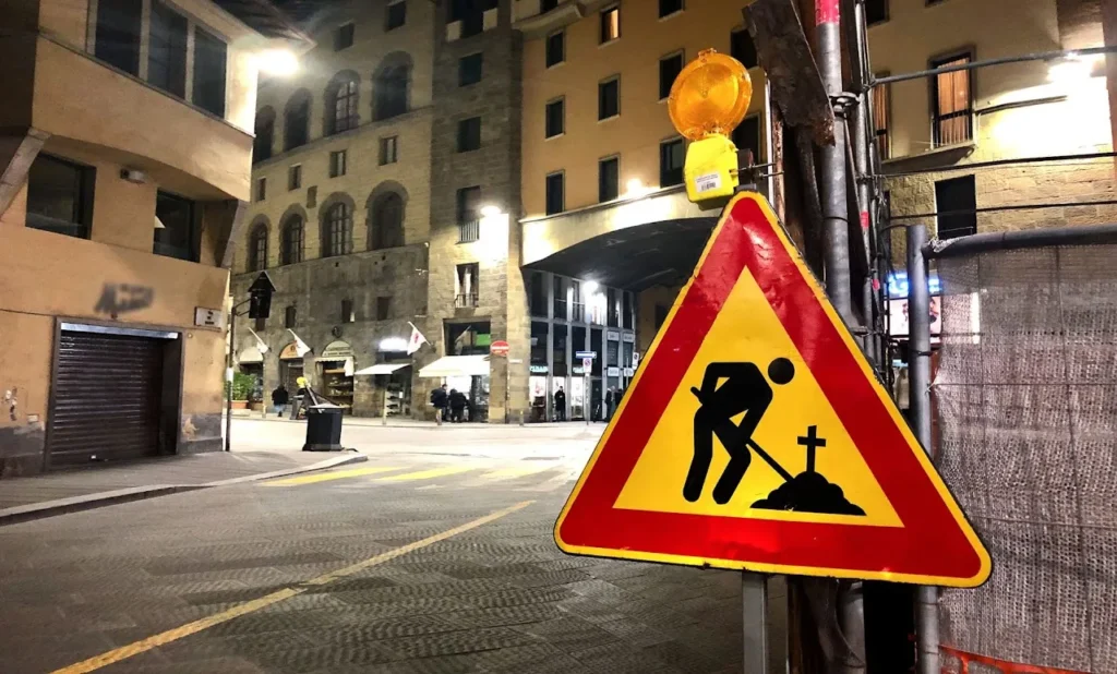 Cover image of the photo gallery for 'Life or Work,' featuring construction signage at a busy intersection with shops and houses in the background, and a worker symbol with a shovel digging into the ground.mound of dirt, with a cross protruding from it. This imagery evokes the notion of work until death, with a touch of dark humor. Amidst the hustle and bustle of daily life, this sign serves as a poignant yet subtly humorous reminder of the transient nature of existence.