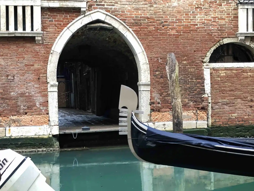 Front view of an artistic installation featuring a brick arch at the entrance, with a gondola passing by. The sea is calm, and the weather is slightly rainy.