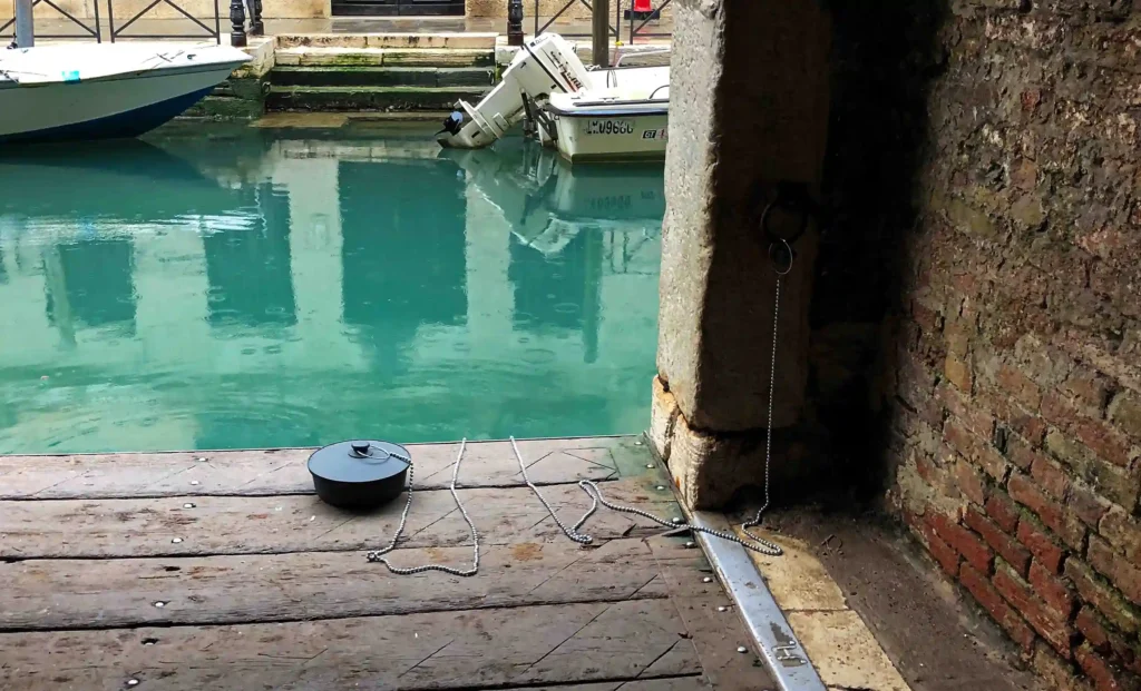 Image from the back of the sculpture, where you can see the entire artistic installation, plus the environment that surrounds it, the plug is on a wooden floor, its chain anchored to the wall, one step away from it it is in water of one of the canals of Venice, and in front of the canal you can see boats that are resting on the other shore, next to walls covered in seaweed, the day becomes a little dark and rainy.