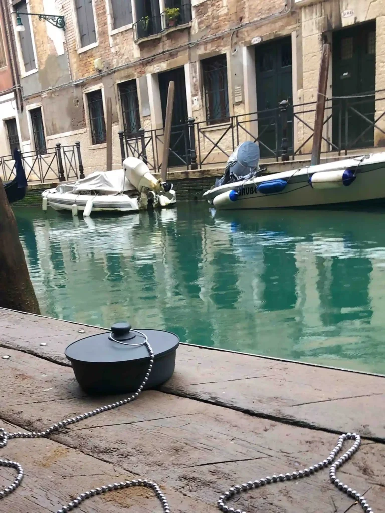 Image of an artistic installation near the canals of Venice, featuring a large drain plug sculpture aimed at raising awareness about global warming. The view is taken from the right corner, showing the wooden floor, calm canal waters, moored boats, and weathered two-story houses in the background.