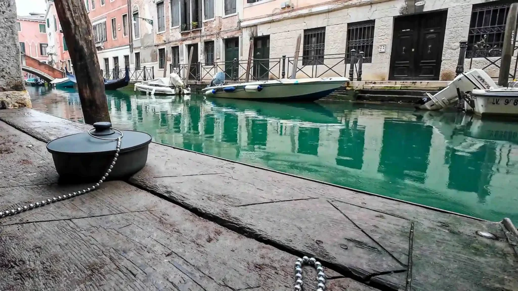 Cover image of the photo gallery for 'Salvezza,' depicting a large drain plug sculpture near the canals of Venice, viewed from the right corner and below, with a worn wooden floor, calm waters, moored boats, and weathered two-story houses in the background.