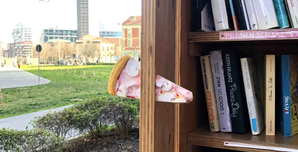 View of a business district with glass buildings on the left and a sculpture of a pizza slice with ham slices marked by euro symbols piercing the wooden door of a street library on the right, symbolizing social equality in everyday eating experiences