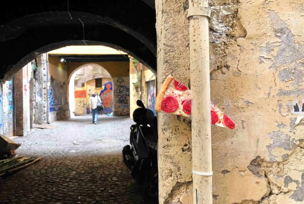 Cover image of the photo gallery for 'Cultural Economy,' showcasing a graffiti-covered alley with a person walking on the left and a pizza sculpture on the right, featuring pepperoni slices engraved with euro symbols, representing the link between urban culture and Italy's economy.