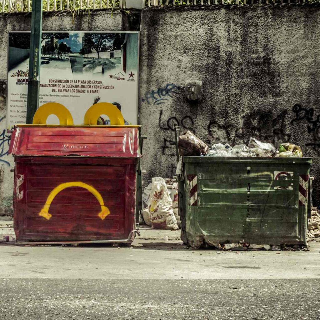 Image of two garbage containers, one painted red with a sad yellow smile and two yellow handles to give the impression that it's a children's menu from a certain fast-food restaurant. Artwork created in Venezuela, a country where many people are forced to eat from the garbage.