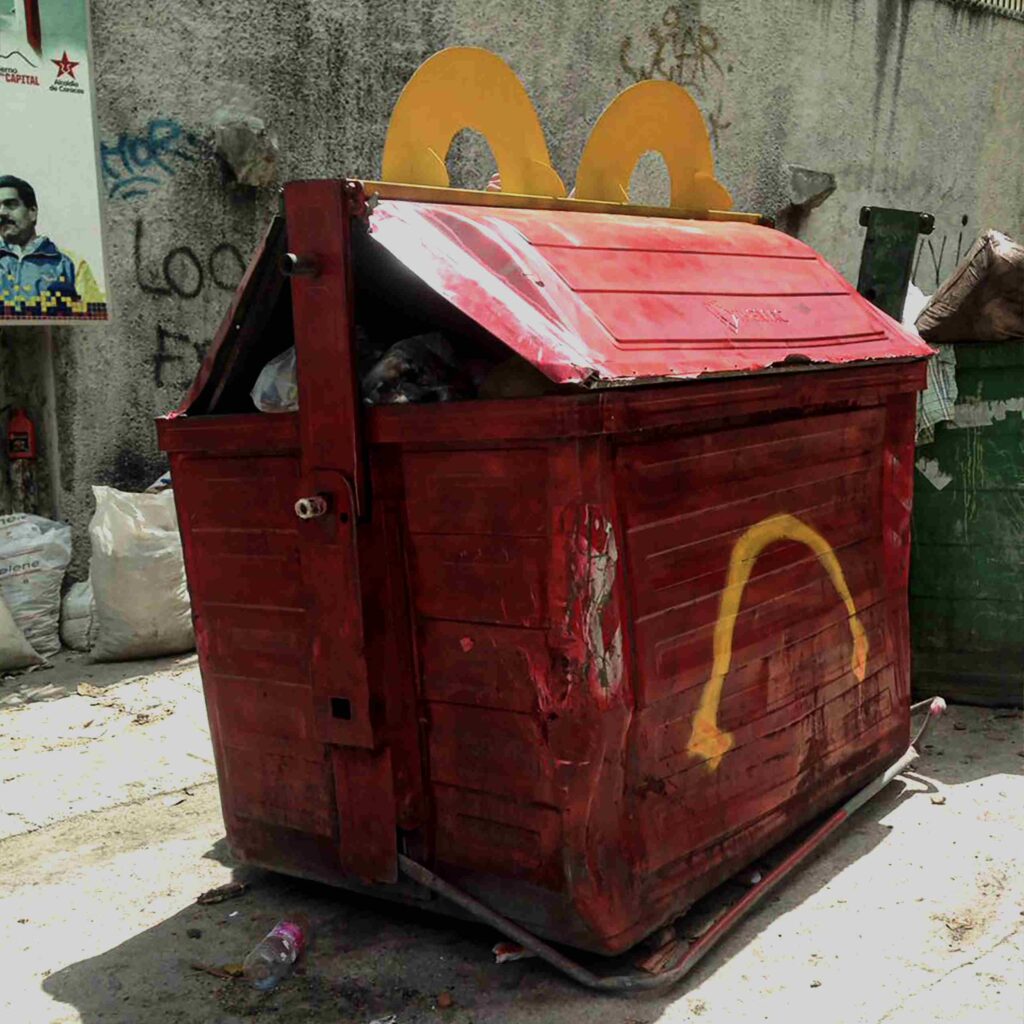 Image of garbage containers, one painted red with a sad yellow smile and two yellow handles to give the impression that it's a children's menu from a certain fast-food restaurant. Artwork created in Venezuela, a country where many people are forced to eat from the garbage.